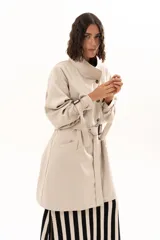 Trench corto color beige con cuello alto, cierre frontal de botones y cinturón ajustable en la cintura. Presenta mangas largas con detalles de tiras ajustables en los puños y bolsillos laterales.