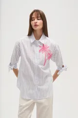Camisa de algodón con rayas verticales azules y blancas, corte ancho y manga larga con lazos ajustables en los puños. Presenta un bordado de dos palmeras en color rosa brillante en el área del pecho.