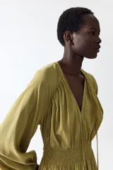 Vestido corto oversized de mezcla de viscosa y color verde militar, con escote en V profundo, cordón ajustable en el cuello y mangas globo largas con puños elásticos. Presenta fruncidos en el canesú y sección de punto smock en la cintura con falda de leve vuelo.