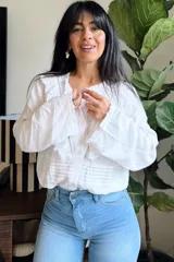 Camisa blanca de estilo romántico con volados en el frente, detalles de encaje y mangas largas con puños elásticos. Presenta un diseño con alforzas en la parte inferior y cierre frontal con botones.