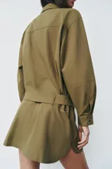 Vestido corto verde militar de cuello solapa y manga larga, con bolsillos de plastrón en el delantero y cierre frontal con cremallera oculta por sopata. Cuenta con un cinturón incorporado del mismo tejido con hebilla metálica y aberturas laterales en el bajo.