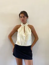 Blusa musculosa de color natural con cuello alto y abertura frontal tipo gota. El diseño permite diversas formas de uso, incluyendo atado al cuello.
