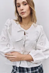 Blusa blanca de tela broderie con diseño de flores caladas, mangas largas abullonadas con puños elásticos y cierre frontal mediante lazos decorativos.