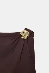 Body negro de manga corta con escote asimétrico, detalle de fornitura metálica dorada en relieve en el hombro y tejido drapeado en el torso. Cierre inferior con botones a presión.