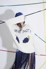 Sweater tejido a mano en lana merino color naranja, con bordado de texto en azul y apliques de uvas en azul. De calce holgado, cuello redondo y puños ajustados.