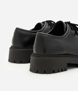 Zapato estilo desert shoe u oxford de Satinato, confeccionado en poliuretano (PU) negro con acabado texturizado. Presenta diseño de punta redonda, cordones al tono y suela gruesa tractorada de goma.