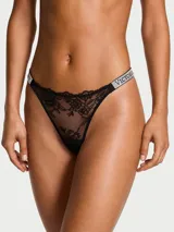 Tanga de encaje negro con tiras laterales brillantes decoradas con pedrería y el logo de la marca.