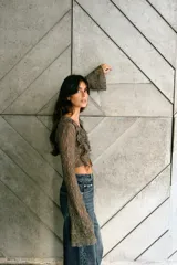 Chaqueta corta de tejido tipo encaje color verde oliva, con escote pico, manga larga acampanada y detalle de volantes en el borde del escote y los puños. Cierre frontal mediante lazadas.