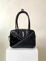 Cartera rígida negra con detalles en rojo y tachas plateadas. Tiene herrajes y cierre metálico plateado marca YKK, asa fina para llevar al hombro o en la mano y correa larga extra regulable. El interior es forrado, tiene un bolsillo con cierre enmarcado en cuero, y un segundo bolsillo plaqué.