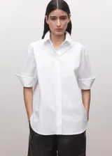 Camisa blanca de algodón con diseño recto, cuello camisero, manga corta y cierre frontal de botones ocultos con tapeta.