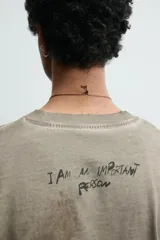 Camiseta de corte relajado con cuello redondo y manga corta. Presenta un efecto de lavado desgastado en tonos marrones y un estampado frontal de una cabeza de alienígena en color verde vibrante.