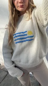 Buzo de punto color crema, tejido a mano en lana merino, con diseño de la bandera de Uruguay bordada en el pecho. Presenta cuello redondo y mangas largas con puños acanalados.