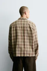 Camisa de corte cropped confeccionada en algodón, con estampado de cuadros en tonos beige, marrón y rojo. Presenta cuello solapa, cierre frontal de botones, manga larga con puño abotonado y detalles de roturas distribuidas por toda la prenda con un bajo irregular.