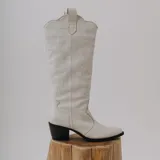 Botas altas estilo texanas de cuero color blanco, con costuras decorativas y bordados en la caña, puntera perfilada y taco ancho oscuro.