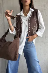 Chaleco color chocolate con bordados en hilo blanco en los bordes y frente. Presenta detalles de tachas metálicas en los hombros y cierre frontal.