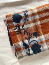 Bufanda de gran tamaño con diseño de cuadros en tonos naranja, beige y azul, decorada con un bordado floral en color azul oscuro y terminaciones con flecos.