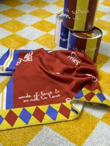Pañuelo cuadrado de seda con diseño estampado en tonos naranja, azul, amarillo y blanco, con logo de la marca.