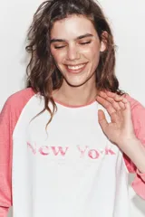 Camiseta de manga larga tipo ranglán con cuerpo blanco y mangas en color rosado. Presenta un estampado frontal con la inscripción 'New York' en tono rosado.