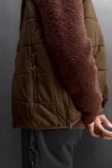 Chaleco marrón acolchado con cuello alto, cierre frontal con cremallera, bolsillos laterales con cremallera y bajo ajustable con trabilla y cremallera lateral.