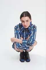 Camisa de corte holgado confeccionada en crepé fino, con estampado geométrico de rayas verticales en tonos azul, blanco y negro. Presenta cuello clásico de tamaño ligeramente ampliado, cierre frontal con botones, mangas largas con pinzas laterales y puños anchos.