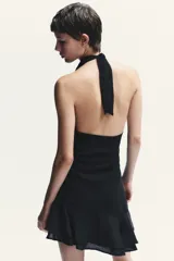 Vestido corto negro de tejido fluido con cuello halter que se ata con lazada. Tiene forro interior, detalle de frunces y bajo asimétrico con volantes. Cierre lateral con cremallera oculta.