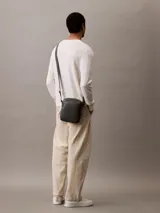 Bolso bandolera Calvin Klein color gris oscuro, de cuero sintético con textura granulada. Tiene correa ajustable, cierre superior con cremallera, bolsillo frontal con cremallera y logo de la marca grabado en la parte inferior.