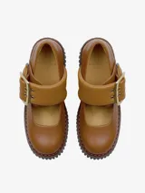 Zapato tipo Mary Jane de cuero color camel, con suela gruesa y dentada color azul marino. Presenta una tira ancha con hebilla metálica dorada y un detalle de ribete azul claro en el lateral de la tira.