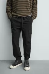 Pantalón cargo de corte slim fit en color negro, confeccionado en tejido de algodón con elasticidad. Presenta cintura elástica ajustable con cordón, bolsillos laterales tipo cargo con solapa y bajo acabado en puño elástico.