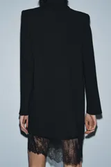 Vestido tipo blazer negro, de corte recto y largo medio, con cuello solapa, manga larga con abertura y botones, y bolsillos con solapa en el delantero. Presenta un bajo con aplicación de encaje negro a tono.