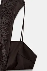 Vestido midi negro de tejido satinado con cuello redondo y manga capa. Presenta detalles de cut-out laterales adornados con encaje a tono. El bajo es asimétrico y tiene cierre en la espalda.