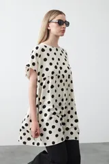 Vestido corto de silueta amplia con corte bajo busto, cuello a la base y mangas cortas. Presenta un estampado de lunares negros sobre fondo beige claro.