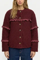 Campera de tela bouclé color bordó, con diseño de cuello redondo y cierre frontal mediante botones. Presenta detalles de volados en tela agamuzada en el pecho y en las mangas.
