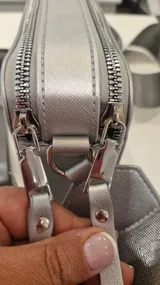 Cartera tipo bandolera de diseño rectangular y estructura rígida, color gris. Cuenta con doble compartimento superior con cierre metálico, herrajes plateados y correa de hombro ancha, ajustable y extraíble.