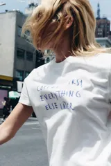 Camiseta blanca de algodón con cuello redondo, manga corta y estampado frontal con el texto en azul "COFFEE FIRST, EVERYTHING ELSE LATER".