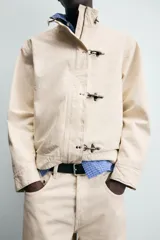 Cazadora de algodón color beige claro, corte relajado, con cuello subido y manga larga. Cierre frontal con cremallera oculta por solapa con cierres metálicos tipo alamar.