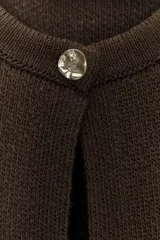 Chaqueta de punto color marrón oscuro, con cuello redondo y manga larga. Presenta flecos decorativos en el bajo y en los puños. Cierre frontal con un único botón dorado.