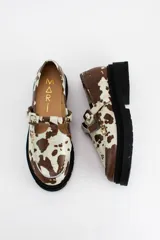 Mocasines estilo Mary Jane con diseño de estampado animal print tipo vaca en tonos marrón y blanco. Presentan una tira con hebilla ajustable sobre el empeine y una suela de plataforma dentada en color negro.