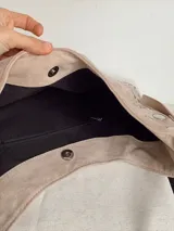Cartera tipo hobo de símil gamuza color beige, con diseño de media luna y decorada con tachas metálicas plateadas en todo su contorno. Incluye correa ajustable y bolsillo interno.