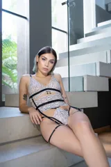 Conjunto de lencería de cuatro piezas compuesto por bralette, tanga, portaligas y ligas. Confeccionado en encaje premium bitono con detalles de tiras elásticas negras y herrajes metálicos. El diseño incluye un bralette con tiras decorativas, tanga de tiro alto con doble tira a la cintura, portaligas desmontable y ligas ajustables.