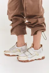 Championes urbanos con diseño de plataforma, combinando paneles en color blanco, dorado y plateado. Presentan cierre mediante tiras de velcro, tirador en el talón y suela dentada en tono marrón.