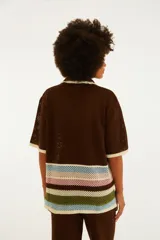 Camisa de punto color marrón con cuello solapa, cierre frontal de botones y mangas cortas. Presenta un bolsillo en el pecho con detalle bordado de una palmera y un tucán, y franjas horizontales multicolor en la parte inferior.