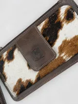 Billetera porta celular confeccionada en cuero marrón con detalle de pelo vacuno con estampado animal print tipo vaca en tonos blancos, marrones y negros. Cuenta con correa de muñeca y cierre superior.