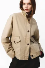 Campera levemente acolchada en lona de algodón gruesa color beige, con cuello y puños vueltos de efecto cuero marrón oscuro. Presenta cierre frontal con botones, solapa rompevientos, bolsillos delanteros de parche con solapa y bolsillos calientamanos.