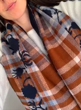 Bufanda de gran tamaño con diseño de cuadros en tonos naranja, beige y azul, decorada con un bordado floral en color azul oscuro y terminaciones con flecos.