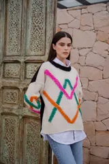 Sweater de punto color crudo con cuello redondo negro y bordados decorativos en forma geométrica en colores fucsia, verde y naranja. Tiene mangas largas con puños negros.