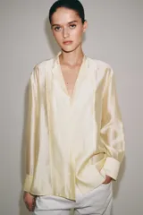 Camisa de mujer fluida de ZARA WOMAN COLLECTION, confeccionada en hilatura con mezcla de lyocell. Presenta escote en pico, manga larga con puños marcados y bajo asimétrico, con cierre frontal de botones ocultos.