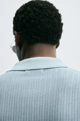 Polo de manga corta, color celeste pálido, con cuello solapa y abertura frontal en V. Tejido de punto con textura acanalada vertical.