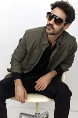 Camisa verde militar de corte recto, con cuello clásico, cierre frontal con botones a presión, mangas largas con puños abotonados, dos bolsillos de parche en el pecho y bajo redondeado.