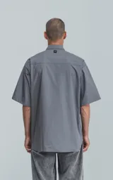 Camisa de manga corta en color gris, confeccionada en popelina de algodón. Presenta un corte oversize, cuello clásico, cierre frontal con botones y dos bolsillos delanteros, uno de ellos con solapa.
