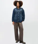 Camisa de jean estilo overshirt con corte oversized, cuello clásico y mangas largas. Presenta cierre frontal con botones, dos bolsillos delanteros con solapa y aplicación de tachuelas metálicas en el cuello y la parte superior del pecho.